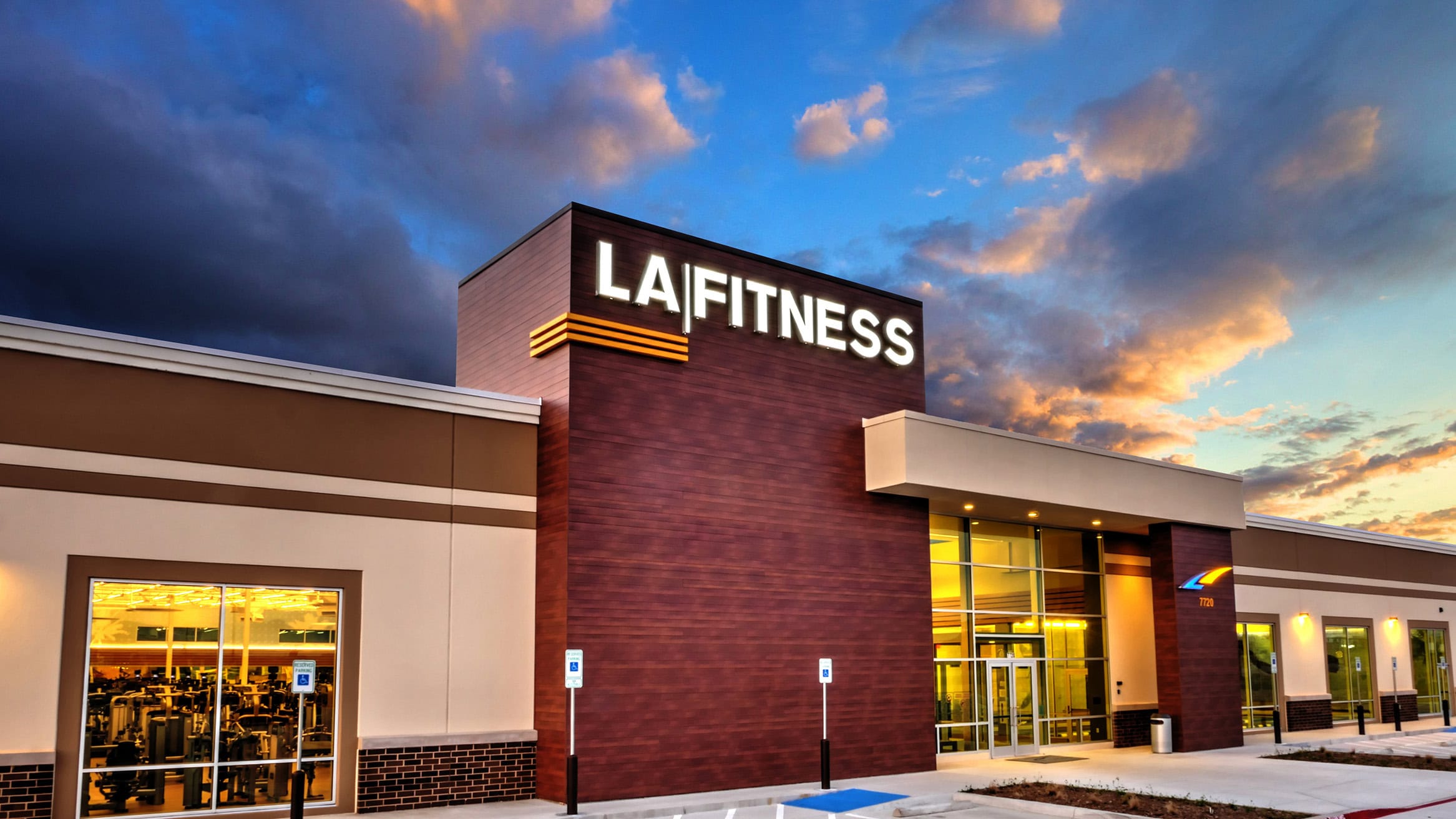 Modern LA Fitness gym exterior at sunset with illuminated signage and exercise equipment visible through large windows, offering a welcoming environment for fitness and health enthusiasts.