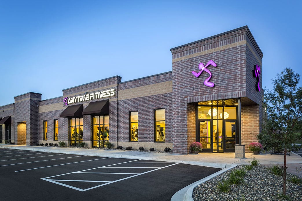 Brightly lit Anytime Fitness gym exterior at dusk in a modern brick building with parking lot; part of Crates Health's fitness and wellness services.